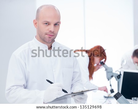close-up of a smiling scientist holding a clip-board in a chemistry lab around a worktable where two other people analyzing under microscope