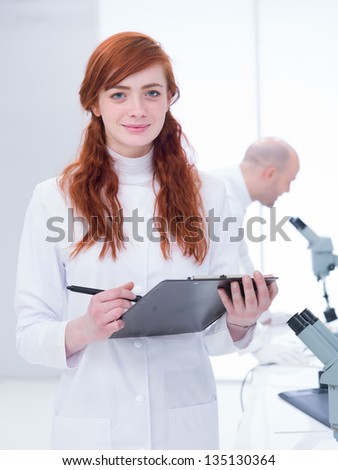 close-up of a smiling student holding a clip-board in a chemistry lab around a worktable with lab tools and her teacher analyzing under microscope in the background