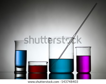 different sizes beakers filled with colored liquid whith a dropper in one, against a gradient gray background and laying on a reflective surface