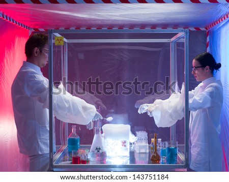 two scientists, a man and a woman, mixing chemicals in a protection enclosure labeled as bio hazardous, into a beaker filled with vapors, in a containment tent