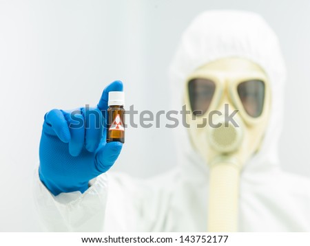Radioactive isotope sample in a small brown bottle with a warning label held in the fingers of a laboratory scientist in full protective clothing with a mask and breathing apparatus