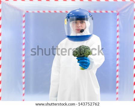 person in protective suit and helmet holding a broccoli with one hand in front of a confinement tent