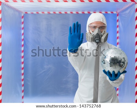 person dressed in a bio hazard protective suit making a halt gesture with one hand and holding a disco ball in the other in front of a confinement tent