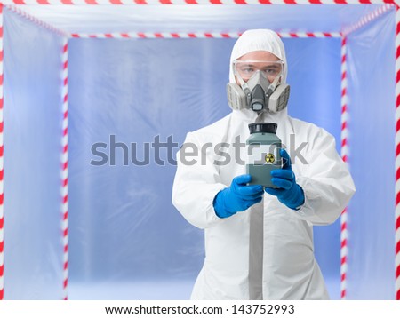 male scientist wearing protection equipment holding a toxic waste container, inside a chamber surounded with red and white tape