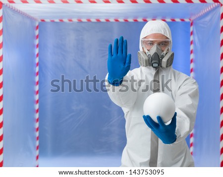 person dressed in a bio hazard protective suit making a halt gesture with one hand and holding a styrofoam sphere in the other in front of a confinement tent