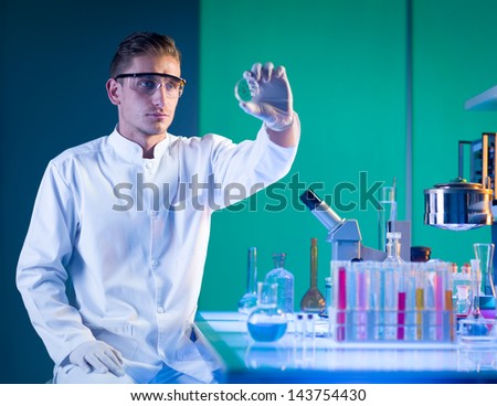 close-up of scientist sitting down near a laboratory table with chemistry glassware on top, holding a petri dish analysing it