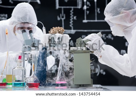 Laboratory scientists in sterile garb wearing masks and safety goggles working at a bench with microscopes surrounded by flasks full of colourful chemicals
