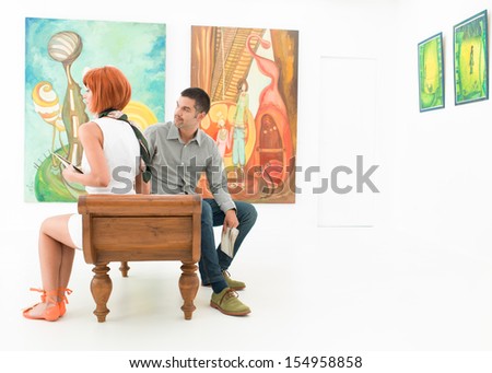 two caucasian people sitting on a wooden bench holding brochures, with colorful paintings on background
