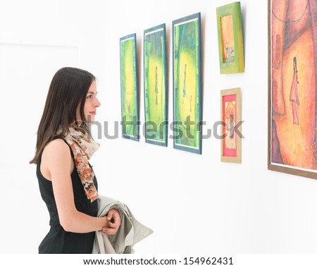 side view of young caucasian woman standing in an art gallery in front of colorful framed paintings displayed on a white wall