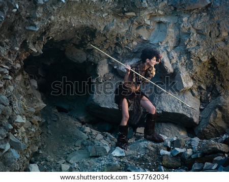 caucasian woman dressed as a caveman holding a spear, getting ready for hunting, close to a rocky cave