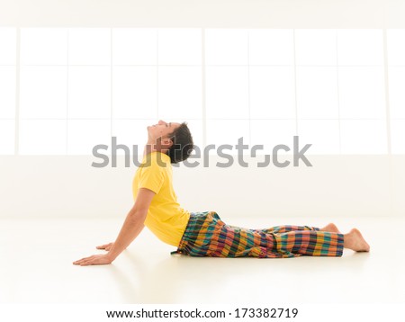 Colorful dressed male repeating bujangasana cobra pose yoga exercises in a white room with window background