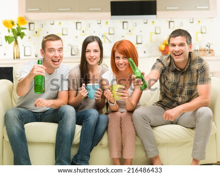 portrait of four young caucasian friends sitting on sofa holding drinks, watching a television show