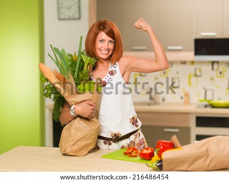 front view of beautiful caucasian woman standing in kitchen, smiling, holding bag with groceries, showing her arm muscles
