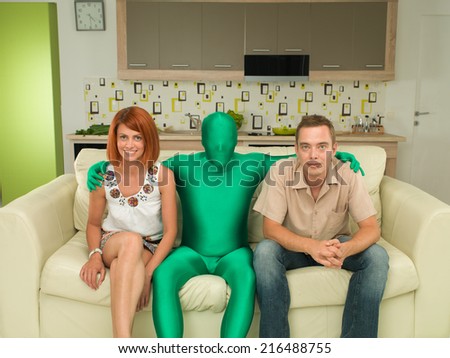 young caucasian man and woman sitting on couch with man dressed in green full body suit between them, hugging