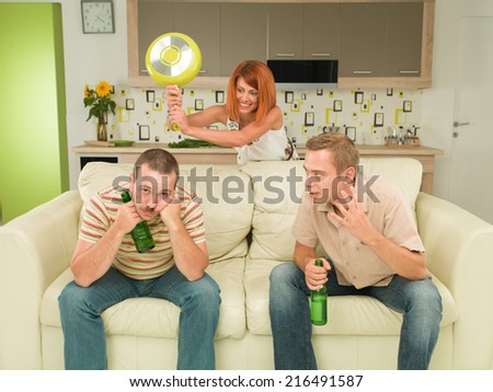 two caucasian men sitting on couch holding beer bottles, having conversation, woman in background trying to hit one of them with frying pan
