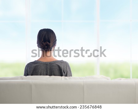 back view of young caucasian woman sitting on couch enjoying the view outside