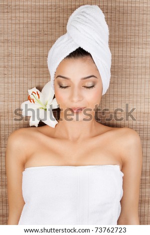 portrait of a beautiful, young woman, at a spa, lying on her back on a bamboo mat, with a towel in her head, smiling, with a lily near her head and her eyes closed.