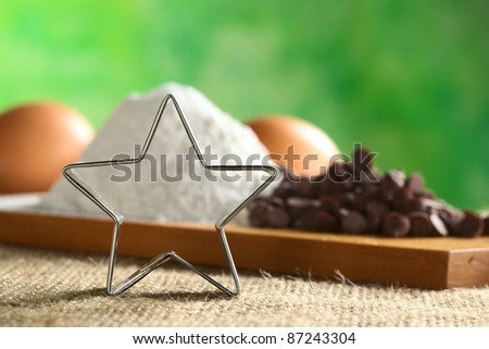 Star-shaped cookie cutter with baking ingredients such as wheat flour, chocolate chips and eggs in the back (Selective Focus, Focus on the cookie cutter)
