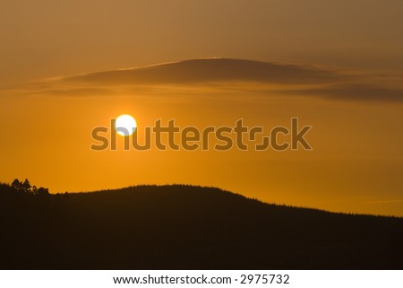 Setting Sun between forested hill and a cloud that mirrors its shape