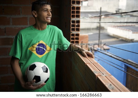 Smiling Brazilian teen wearing Brazilian flag t-shirt stands holding football soccer ball looking out the favela window