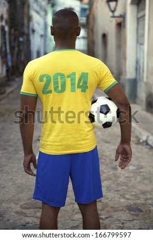 Brazilian football player in 2014 shirt stands holding a soccer ball on an old rustic village street