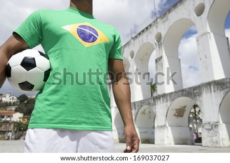 Brazilian football player holding soccer ball wearing Brazilian flag shirt at Lapa Arches in Rio de Janeiro