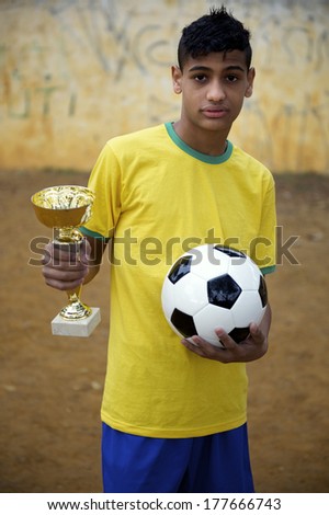 Serious young Brazilian football player holding trophy and soccer ball on basic dirt football pitch