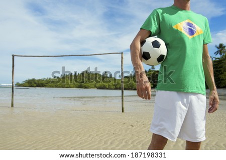 Football player holding soccer ball sitting on simple Brazilian beach football pitch with rustic goal posts sticking out of the sand