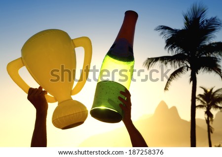 Celebrating hands holding inflatable trophy and champagne bottle above sunset skyline of Rio de Janeiro Brazil