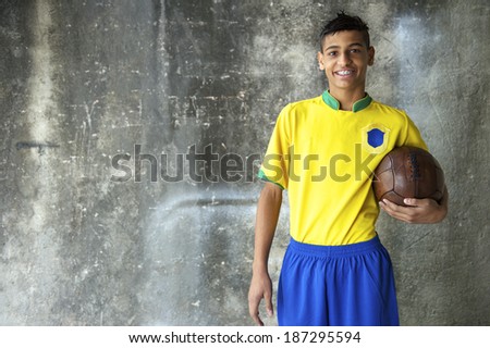Smiling young Brazilian soccer player in team uniform of Brazil colors holding vintage football against concrete favela wall
