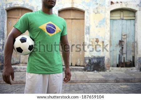 Brazilian football player stands holding soccer ball on an old rustic village street