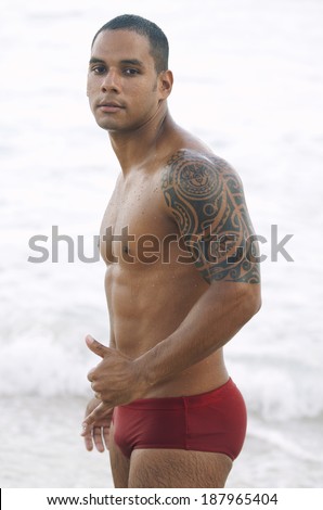 Brazilian man with shoulder tattoo standing on the beach in red sunga bathing suit giving thumbs up Rio de Janeiro