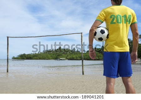 Football player in 2014 shirt holding soccer ball standing on simple Brazilian beach football pitch with rustic goal posts sticking out of the sand