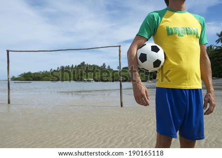 Football player in Brasil shirt holding soccer ball standing on simple Brazilian beach football pitch with rustic goal posts sticking out of the sand