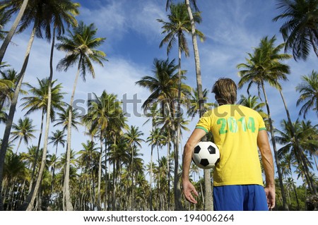 Brazilian football player in 2014 shirt standing in grove of tropical palm trees holding soccer ball Brazil