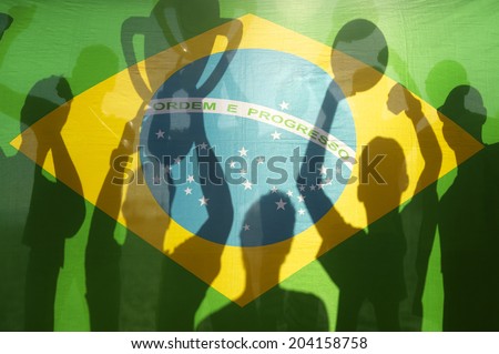 Shadow silhouettes of football team celebrating holding winning trophy and football against Brazilian flag in bright sunlight