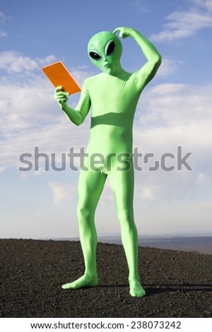 Confused green alien scratching head looking down at orange tablet