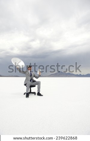 Businessman sitting outdoors in dramatic desert landscape holding satellite communication receiver and digital tablet