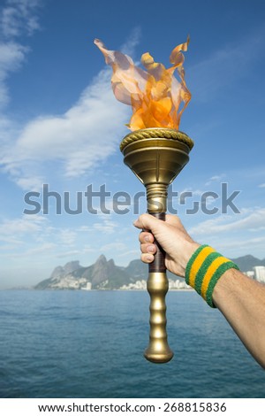Hand of an athlete wearing Brazil colors sweatband holding Olympic sport torch against Rio de Janeiro Brazil skyline with Two Brothers Mountain
