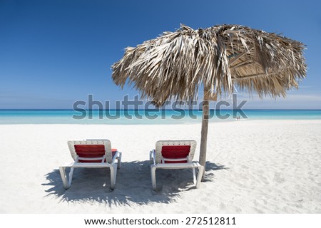 Pair of empty beach chairs sit under palm frond palapa umbrella on an empty stretch of bright Caribbean beach in Varadero Cuba