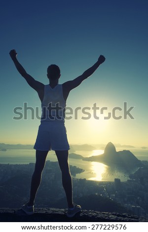 Silhouette of athlete in white sport uniform standing with champion arms raised in front of Rio de Janeiro Brazil sunrise skyline overlook at Sugarloaf Mountain