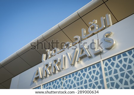 Arrivals sign at an airport in the Middle East features Arabic text and Islamic patterns under modern architecture and bright blue sky