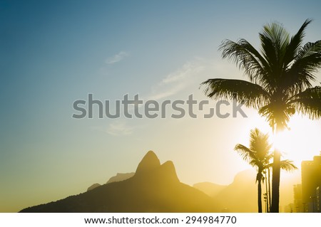 Sunset in Rio de Janeiro Ipanema Beach Brazil with Two Brothers Dois Irmaos Mountain and golden sun through palm trees silhouettes