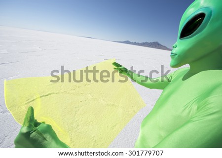 Green alien traveler in white desert lunar landscape reading electronic map on future technology flexible display tablet