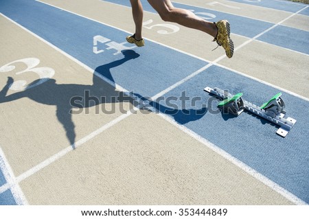 Athlete in gold shoes sprinting from the starting blocks over the starting line of a race on a blue and tan running track