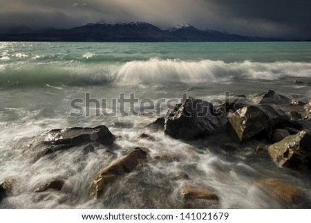 Waves crashing on rocky shore of Lake Pukaki, with storm clouds hanging over the mountains. New Zealand
