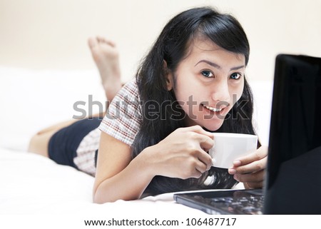 Smiling sweet asian woman with laptop and cup of coffee on the bed