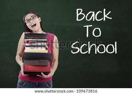 Portrait of female high school student holding pile of textbooks with text of Back To School on the blackboard