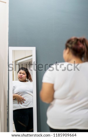 Portrait of obese woman looking at her fat belly while standing near a mirror at home