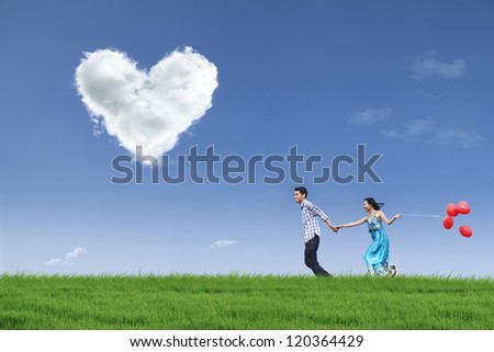 Happy couple is running together in green field while holding red balloons